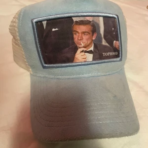 Blå james bond keps från Tophood - Snygg blå keps från Tophood med ett retrotryck på framsidan. Kepsen har en mesh-baksida för bättre ventilation och justerbar passform. Perfekt för sommaren! Hör av er vid funderingar:)