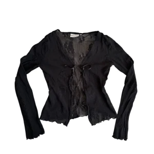 Svart mesh/blus med knytning - Superfin svart långärmad topp i tunn, mesh-liknande spets med snygga detaljer. Har en feminin passform med knytning framtill och vågiga kanter för en romantisk och vintage-inspirerad look. Perfekt att styla över ett linne eller en spets-bh!