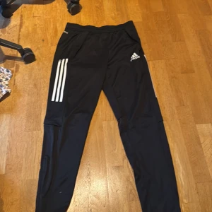 Svarta träningsbyxor från Adidas - Säljer ett par svarta träningsbyxor från Adidas med vita ränder på sidan och logotyp på låret. Byxorna har en elastisk midja och dragkedjor vid anklarna för enkel på- och avtagning. Perfekta för träning eller en avslappnad dag.