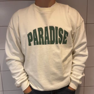 Vit sweatshirt med texten 'PARADISE' - Säljer en snygg vit sweatshirt med grön text 'PARADISE' på framsidan. Tröjan är långärmad och har en normal passform. Perfekt för en avslappnad stil och passar till alla tillfällen. Superbekväm och stilren!