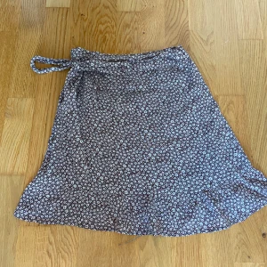 Blommig omlottkjol - Säljer en superfin blommig omlottkjol med volangdetaljer. Kjolen är perfekt för sommardagar och har en söt knytning i midjan. Använt 1-2 gånger så den är i nyskick!