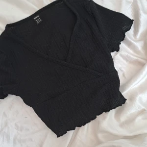 Svart omlottopp från Shein - Säljer en svart omlottopp från Shein i storlek XS. Toppen har en v-ringad design och korta ärmar. Materialet är lätt och luftigt, perfekt för en sommarkväll. Den har en croppad passform som ger en trendig look.