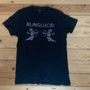 Svart t-shirt med blingdetaljer - Snygg svart t-shirt från Blinglucid med glittriga detaljer i form av två änglar och texten 'BLINGLUCID' på framsidan. Perfekt för dig som vill sticka ut med lite extra bling!