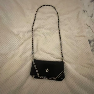 Svart axelväska med kedjedetaljer - Snygg svart axelväska med en stjärndetalj på framsidan. Väskan har en kedja som axelrem och dragkedja upptill. Perfekt för att ge en edgy touch till din outfit.