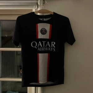  PSG fotbollströja - Snygg svart PSG-tröja med Mbappé 7 på ryggen. Framsidan har Qatar Airways-logga och PSG-emblem. Kortärmad design med vit och röd detalj på framsidan. Perfekt för fotbollsfans!