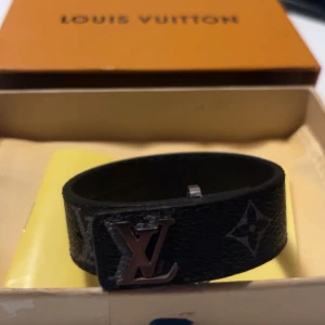 Svart armband från Louis Vuitton - Elegant svart armband från Louis Vuitton med det ikoniska LV-emblemet. Armbandet har en stilren design med monogramdetaljer och är perfekt för att ge en lyxig touch till din stil.
