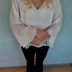 Vit blus med v-ringning - Elegant vit blus med v-ringning och långa ärmar. Blusen har en lös passform och är tillverkad i ett lätt och luftigt material, perfekt för både vardag och festliga tillfällen. Storlek: passar både xs och s. 