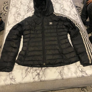 Svart dunjacka från Adidas - Säljer en svart dunjacka från Adidas med huva och dragkedja. Jackan har de klassiska Adidas-ränderna längs ärmarna och en broderad logga på bröstet. Perfekt för kyliga dagar! Och den har några hål som är bytte liten