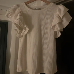 Vit ribbad topp från Zara - Säljer en stilren vit ribbad topp från Zara med volangdetaljer på ärmarna. Perfekt till en sommardag!