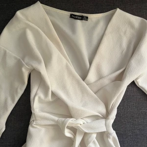 Vit omlottopp från Boohoo - Snygg vit omlottopp från Boohoo med knytband i midjan. Toppen har en v-ringad design och långa ärmar, perfekt för en stilren look.