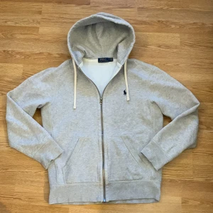 Grå hoodie från Polo Ralph Lauren - Säljer en stilren grå hoodie från Polo Ralph Lauren med dragkedja och broderad logga på bröstet. Tröjan har långa ärmar och en praktisk huva med snörning. Nyskick