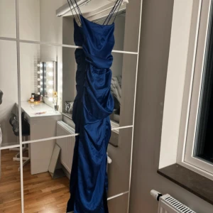 Blå balklänning  - Elegant blå långklänning med smala axelband och volangdetaljer längs sidorna. Perfekt för en festlig kväll med sin stilrena design och vackra fall.