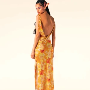 Gul blommig långklänning - Elegant gul långklänning med blommigt mönster och öppen rygg. Klänningen har smala axelband och är tillverkad i ett glansigt material som ger en lyxig känsla och en slits. Helt ny, oanvänd.