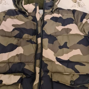 Camouflagefärgad pufferjacka från Lindex - Säljer en vattenavvisande pufferjacka från Lindex i camouflagefärger. Jackan har en huva och dragkedja framtill, samt praktiska fickor med knappar. Perfekt för kyliga dagar.