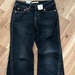 Svarta jeans med slitningar - Snygga svarta Levi's jeans med slitningar på knäna och kontrasterande sömmar. Klassisk femficksdesign med orangea nitar och en läderpatch baktill. Perfekta för en avslappnad stil.