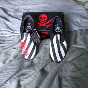 Svartvita slip-ons från Draven - Aldrig använt. Helt nya. Unika slip-ons från Draven med svartvitt randigt mönster och röd dödskalle. Skorna har en klassisk slip-on design med elastiska sidor för enkel på- och avtagning. Perfekta för en edgy stil.