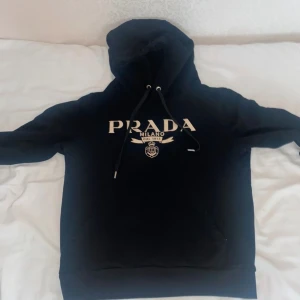Svart hoodie från Prada - Snygg svart hoodie från Prada med vit logga framtill. Perfekt för en stilren och bekväm look. Hoodien har en klassisk design med dragsko i huvan och ribbade muddar vid ärmslut och nederkant. Pris kan diskuteras 