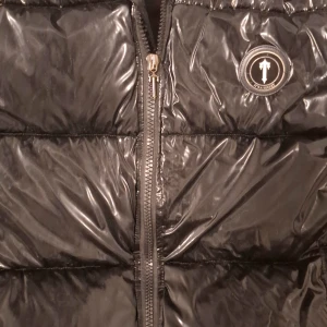 Svart pufferjacka från Trapstar - Snygg svart pufferjacka från Trapstar med glansigt material och dragkedja framtill. Jackan har en stilren design med ett märke på bröstet. Perfekt för kyligare dagar den har ett litet hål i högra sidan vid armen men man märker intr direkt 