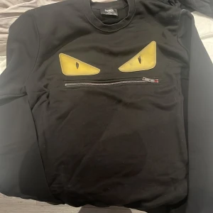 Svart tröja från Fendi med ögonmotiv - Säljer en svart tröja från Fendi med ett coolt ögonmotiv i gult på framsidan. Tröjan har en dragkedja under motivet och är långärmad. Perfekt för en stilren och unik look.