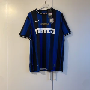 Blå och svart randig fotbollströja från Nike - Säljer en blå och svart randig fotbollströja från Nike med korta ärmar. Tröjan har ett tryck av Pirelli på framsidan och klubbmärke på bröstet. Perfekt för fotbollsfans!retro Champions league tröja Inter 2010