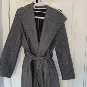 Grå kappa med huva - Snygg grå kappa med huva och knytbälte i midjan. Perfekt för kyliga dagar med sin långa design och stilrena look. Passar bra till både vardag och mer uppklädda tillfällen.