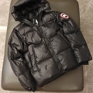 Svart dunjacka från Canada Goose - Snygg och varm svart dunjacka från Canada Goose med huva och dragkedja. Jackan har en klassisk pufferdesign och är perfekt för kalla vinterdagar. Märkets logga syns tydligt på ärmen.