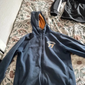 Blå fleecejacka från Sail Racing - Säljer en blå fleecejacka från Sail Racing med dragkedja och huva. Jackan har en broderad logga på bröstet och orange foder. Perfekt för kyliga dagar.