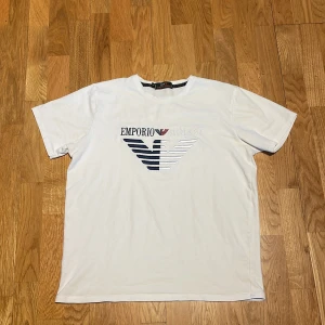 Vit t-shirt från Emporio Armani - Snygg vit t-shirt från Emporio Armani med ett stort broderat logotypmotiv i blått och vitt på bröstet. Perfekt för en stilren och avslappnad look.