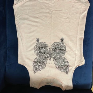 Beige t-shirt med fjärilsmönster från R Sister - Säljer en beige t-shirt från R Sister med ett detaljerat svart fjärilsmönster på framsidan. T-shirten har en rund halsringning och korta ärmar. Perfekt för en avslappnad stil.