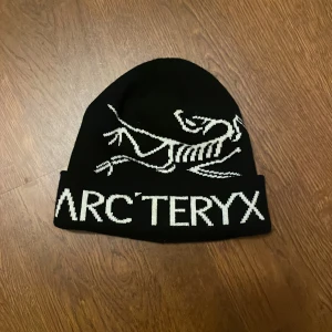 Svart mössa från Arc'teryx - Snygg svart mössa från Arc'teryx med vit logotyp och text framtill. Tillverkad i 100% bomull, perfekt för kyliga dagar. Enkel och stilren design som passar till det mesta.