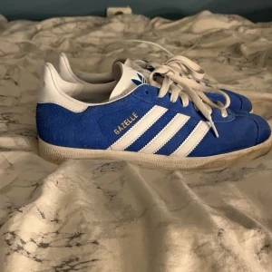 Blå Adidas Gazelle sneakers - Snygga blå Adidas Gazelle sneakers med vita detaljer och snörning. Skorna har den klassiska tre-randiga designen och är tillverkade i mocka. Perfekta för en sportig och stilren look.