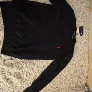 Svart tröja från Polo Ralph Lauren - Snygg svart tröja från Polo Ralph Lauren med det klassiska röda logotypbroderiet på bröstet. Tröjan har långa ärmar och en rund halsringning. Perfekt för en stilren look.