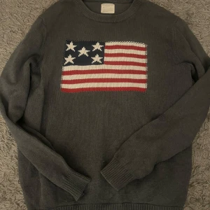 Grå stickad tröja från Selected Homme - Snygg grå stickad tröja från Selected Homme med ett coolt amerikanskt flaggmotiv på framsidan. Tröjan har långa ärmar och ribbade muddar vid ärmslut och nederkant. Perfekt för en avslappnad stil. det står på tröjan att det är storlek XL men den sitter som L/M. skriv vid minsta fundering!!
