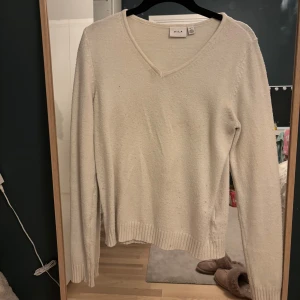 Vit stickad tröja från Vila - Säljer en stilren vit stickad tröja från Vila med v-ringning. Tröjan har långa ärmar och ribbade kanter vid ärmslut. En fläck där uppe vid v-ringningen.