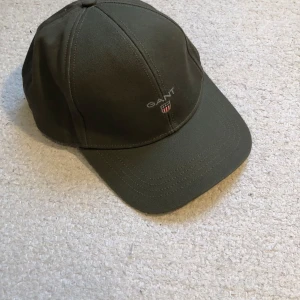 Olivgrön keps från GANT - Snygg olivgrön keps från GANT med broderad logga framtill. Perfekt för en avslappnad stil och skydd mot solen. Justerbar passform för optimal komfort. Nypris: 500kr