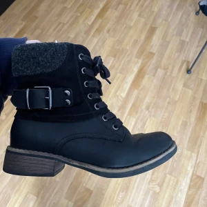 Svarta boots med snörning och bälte - Snygga svarta boots med snörning och dekorativt bälte. De har en robust sula och en bekväm passform. Ovandelen är i skinn med en mjuk textilkrage.