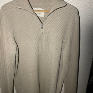 Beige stickad tröja från Jack & Jones - Snygg beige stickad tröja från Jack & Jones med dragkedja i halsen. Perfekt för kyligare dagar med sin ribbade struktur och långa ärmar. En stilren och bekväm tröja för alla tillfällen.