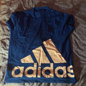 Blå t-shirt från Adidas - Snygg blå t-shirt från Adidas med det klassiska logotyptrycket i vitt på framsidan. Perfekt för en sportig look eller avslappnad stil. Kortärmad och bekväm passform.