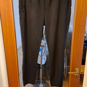 Svarta byxor med sidodetaljer - Snygga svarta byxor med diskreta sidodetaljer. Perfekta för en stilren look. Byxorna rekommenderas till personer med kortare ben. tillverkade i ett bekvämt material. Finns några lösa trådar här och där.