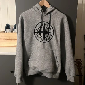 Grå hoodie från Stone Island - Säljer en stilren grå hoodie från Stone Island med det ikoniska märket på ärmen. Tröjan har en stor logga framtill och en praktisk känguruficka. Perfekt för en avslappnad look. Säljer eftersom jag har vuxit ur den. 