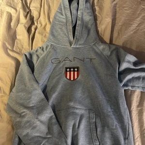 Ljusblå hoodie från GANT - Säljer en snygg ljusblå hoodie från GANT med broderad logga och emblem på bröstet. Tröjan har en klassisk känguruficka och en bekväm huva. Perfekt för en avslappnad stil.