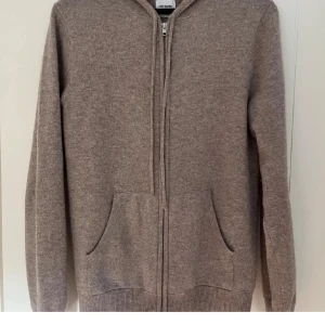 Beige cashmere hoodie från John Henric - Säljer en lyxig beige hoodie i mjuk cashmere från John Henric. Tröjan har dragkedja och praktiska fickor framtill. Perfekt för kyliga dagar med sin stilrena design och bekväma passform.