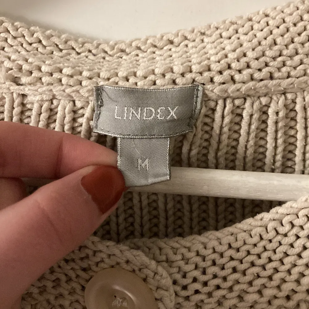 Säljer en stilren beige stickad kofta från Lindex med stora knappar framtill. Koftan har en ribbad design och långa ärmar, perfekt för kyliga dagar. Passar utmärkt till både vardag och lite finare tillfällen.. Neuletakit & Villapaidat.