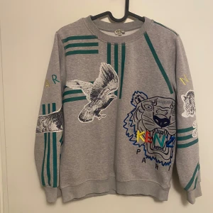 Grå sweatshirt från Kenzo med djurmotiv - Säljer en grå sweatshirt från Kenzo med coola djurmotiv som tiger, lejon och elefant. Tröjan har gröna ränder och broderade detaljer i flera färger. Perfekt för dig som vill sticka ut!