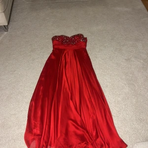 Röd axelbandslös klänning från Porium - Elegant röd axelbandslös klänning från Porium med glittriga detaljer på bysten. Klänningen har en vacker, flytande långkjol och en dragkedja i ryggen. Perfekt för speciella tillfällen.