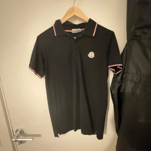 Svart pikétröja från Moncler - Snygg svart pikétröja från Moncler med klassisk krage och röd-vit randig detalj. Tröjan har en broderad Moncler-logga på bröstet och knappar framtill. Perfekt för en stilren look.