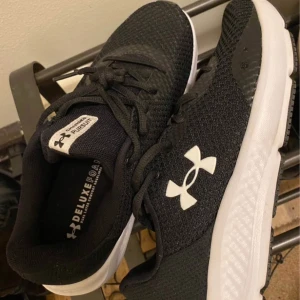 Svarta sneakers från Under Armour - Snygga svarta sneakers från Under Armour med vit sula och klassisk logga på sidan. Skorna har snörning och är perfekta för både träning och vardag. Bekväma och stilrena med en sportig look.