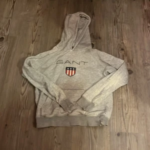 Grå hoodie från GANT - Säljer en stilren grå hoodie från GANT med deras ikoniska logga och ett amerikanskt emblem på bröstet. Tröjan har en klassisk passform med långa ärmar och en bekväm huva. Perfekt för en avslappnad stil.