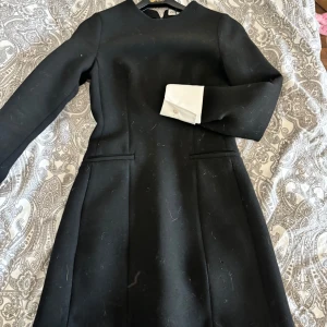 Svart klänning med vita detaljer - Elegant svart klänning med långa ärmar och vita manschetter. Perfekt för en stilren look. Klänningen har en klassisk design med en figurnära passform.