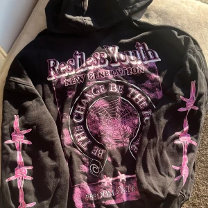 Svart hoodie med tryck - Cool svart hoodie med tryck i rosa och lila. Texten 'Restless Youth' och 'The Change Be The Future' pryder framsidan tillsammans med ett spindelnätsmotiv. Ärmarna har rosa taggtrådsmönster. Perfekt för en avslappnad stil.